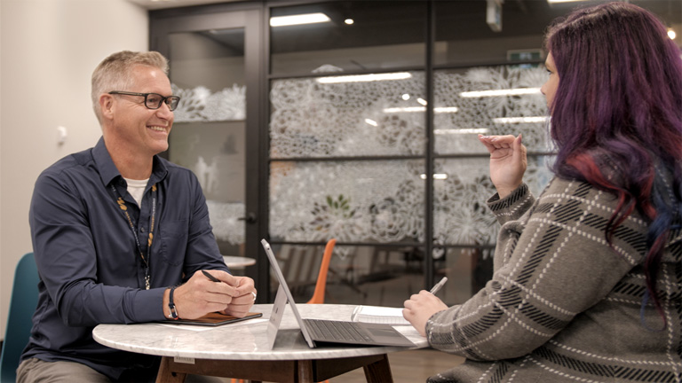 Two employees from Mercury Filmworks sit and talk at a table together as one smiles.
